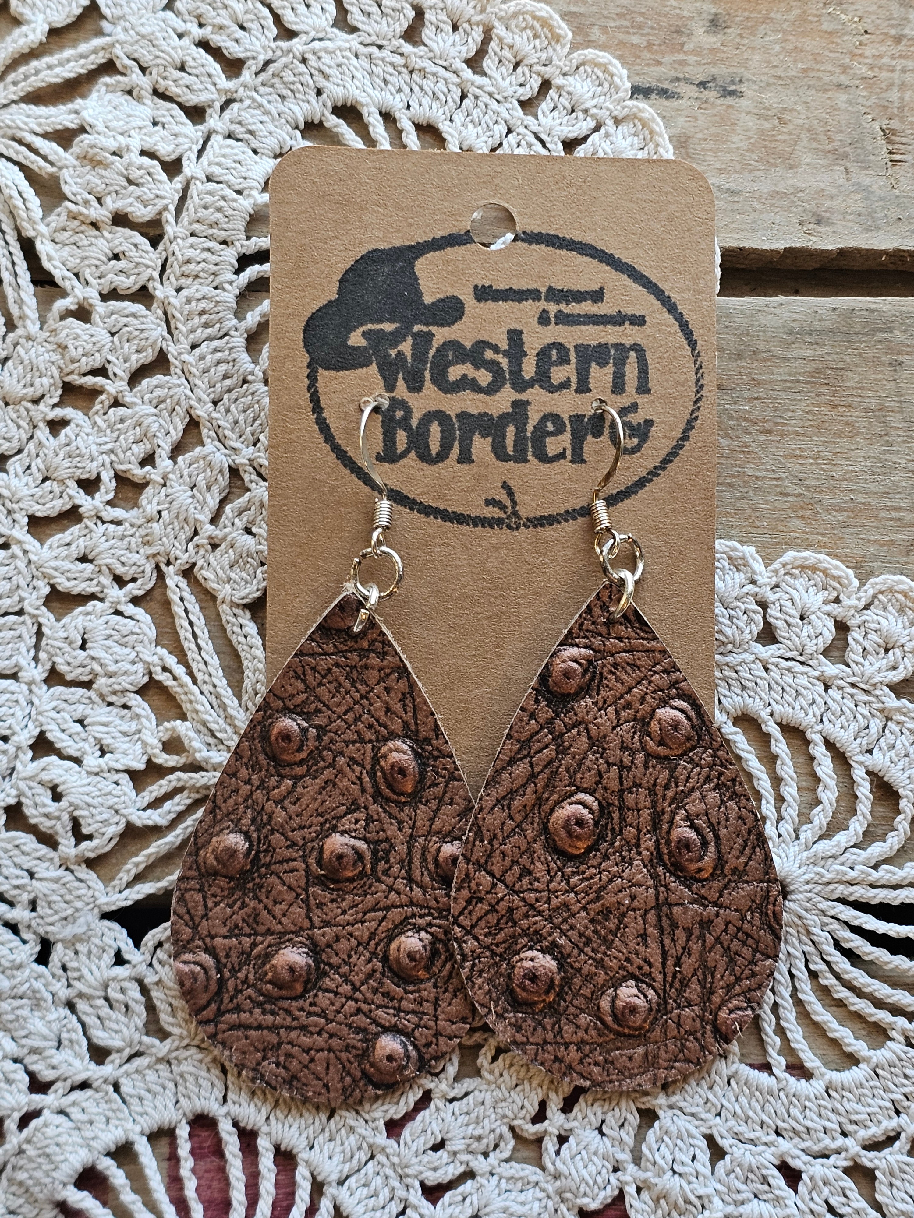 Heart-shaped leather ornament with studded details on a 'Western Border' branded card, placed on a textured fabric background.