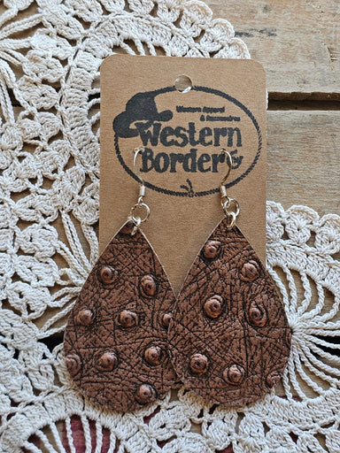 Heart-shaped leather ornament with studded details on a 'Western Border' branded card, placed on a textured fabric background.